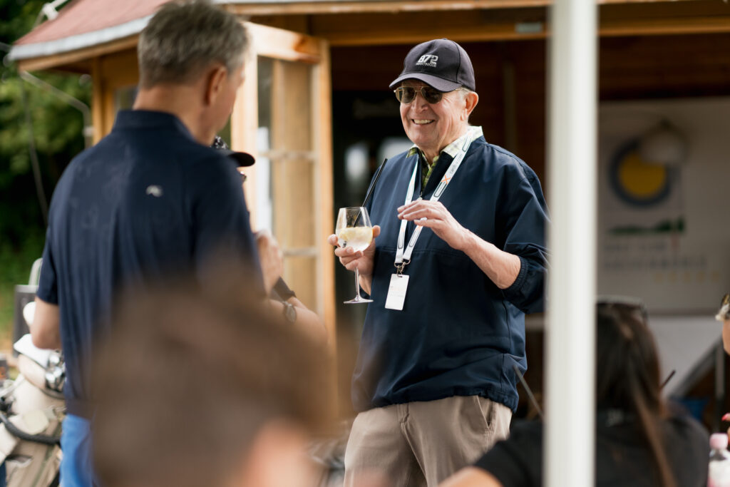 Content Fotograf, ein älterer Herr, mit einem Glas Wein in der Hand. Er lächelt und unterhält sich mit einem anderen Herren.