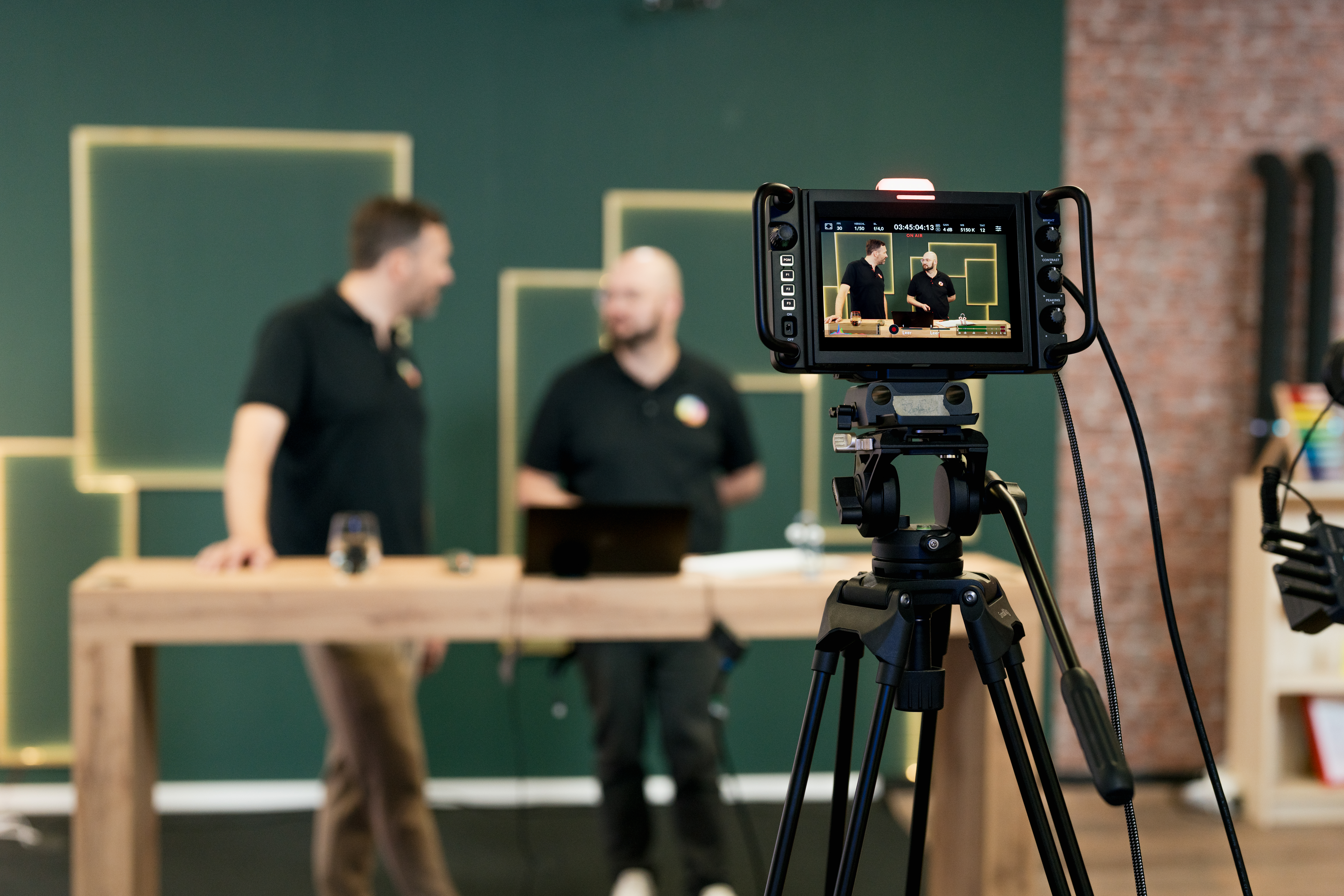 Content Fotograf, Zwei Männer im Hintergrund beide unterhalten sich an einem Tisch, man erkennt sie nur unscharf. Im Vordergrund steht eine Kamera, die auf die beiden gerichtet ist.