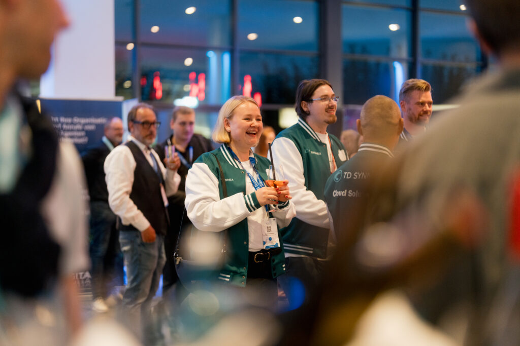 Content Fotograf, Menschen auf einer Messe. Im Vordergrund sind eine Frau und ein Herr mit College Jacken, die einem Vortrag lauschen und einen Aperol trinken.