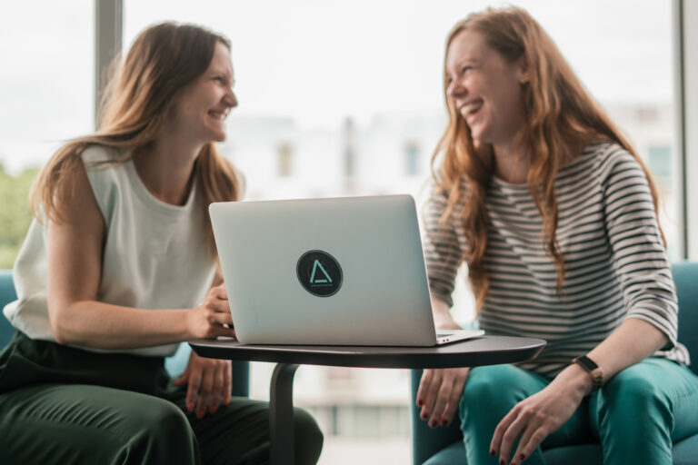 Zwei Frauen sitzen gemeinsam an einem Laptop und sprechen miteinander.