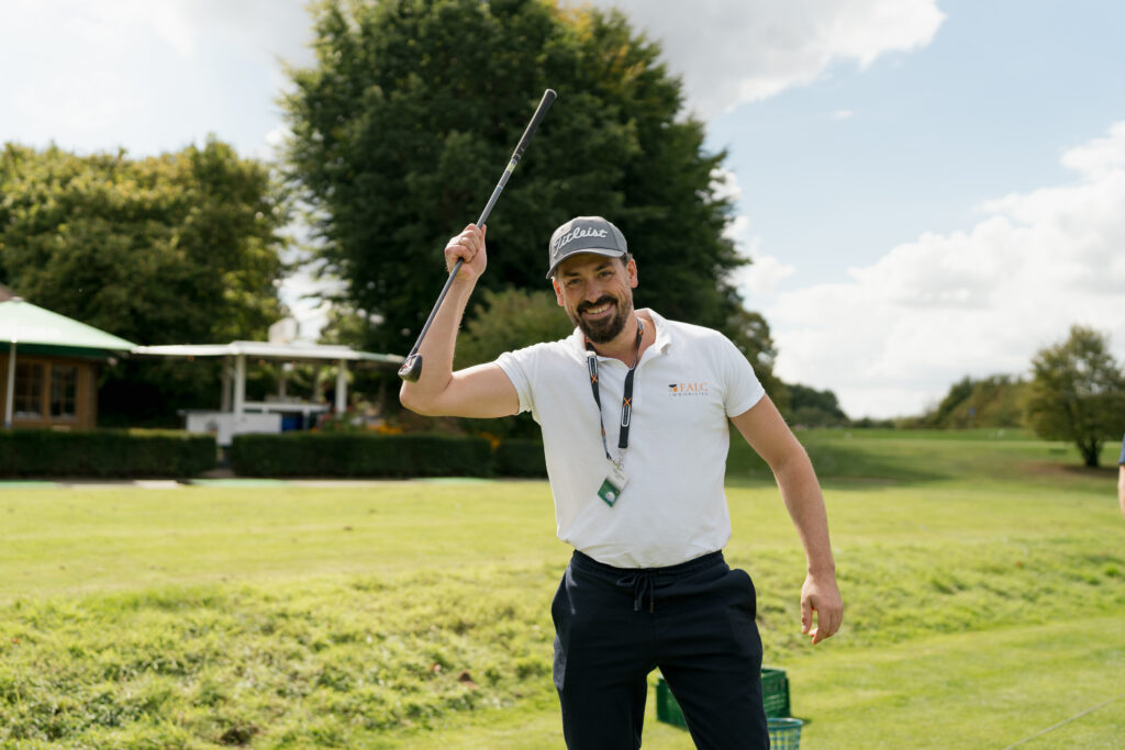 Content Fotograf, lächelnder Mann mit Sportoutfit und einem Golfschläger on der linken Hand. Er steht auf einer Wiese.