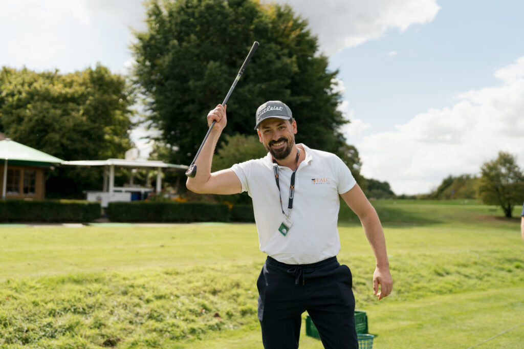 Fototermin Ablauf: Was Dich bei einem Business-Shooting wirklich erwartet 10 Content Fotograf, lächelnder Mann mit Sportoutfit und einem Golfschläger on der linken Hand. Er steht auf einer Wiese. - Fototermin Ablauf