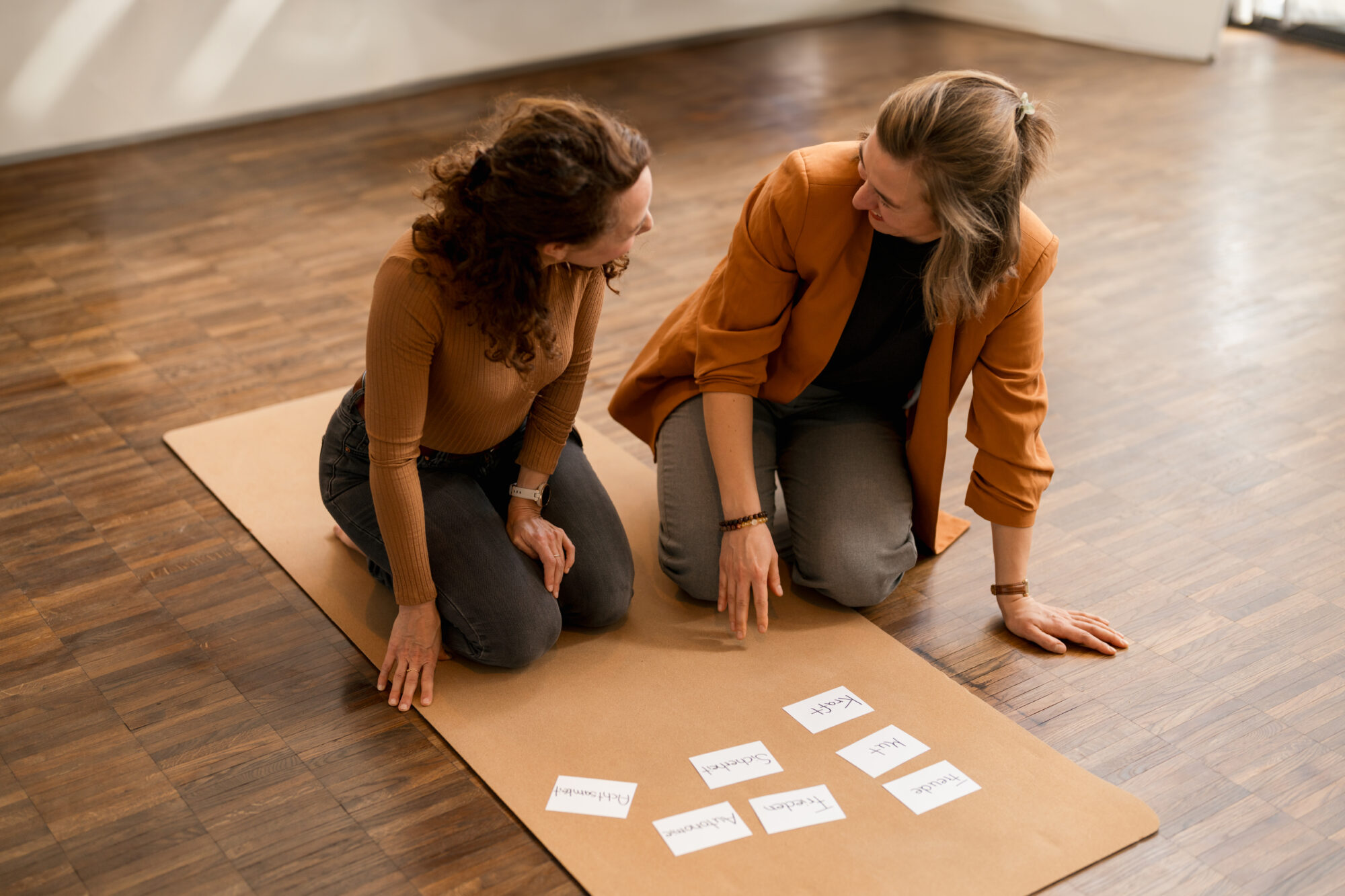 Zwei Frauen sitzen kniend auf einer Yogamatte und planen etwas mit Karteikarten.